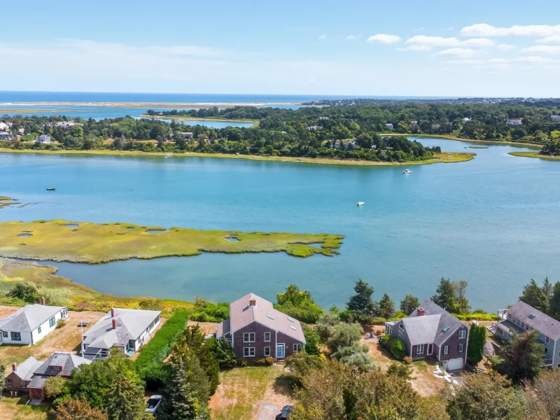 Aerial view of a Cape Cod landscape featuring waterfront homes, lush greenery, and vibrant blue water under a clear sky.