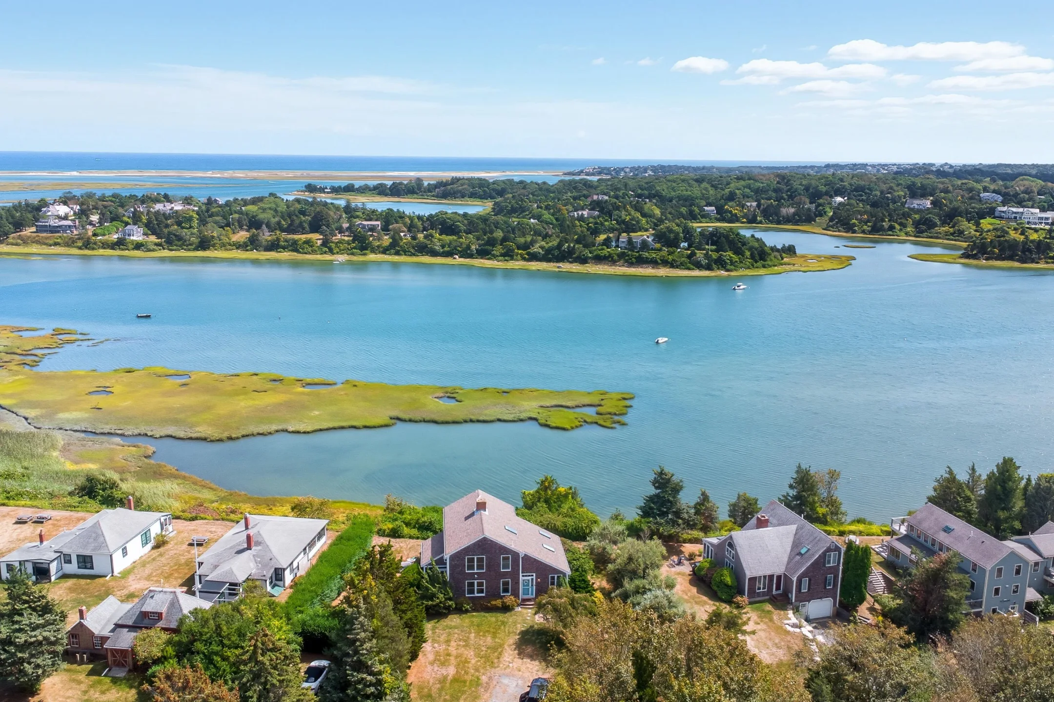 Aerial view of a Cape Cod landscape featuring waterfront homes, lush greenery, and vibrant blue water under a clear sky.