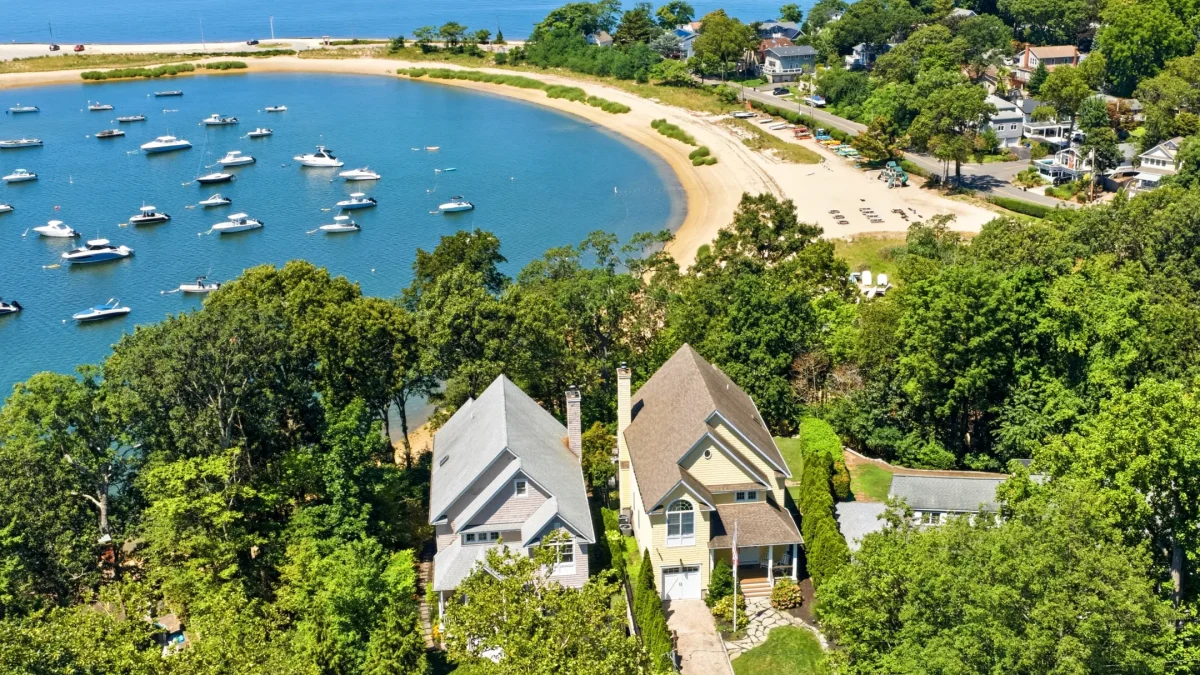 Aerial view of two modern homes surrounded by lush greenery, overlooking a calm bay filled with boats and a sandy beach.