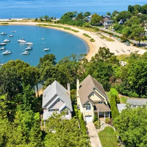 Aerial view of two modern homes surrounded by lush greenery, overlooking a calm bay filled with boats and a sandy beach.