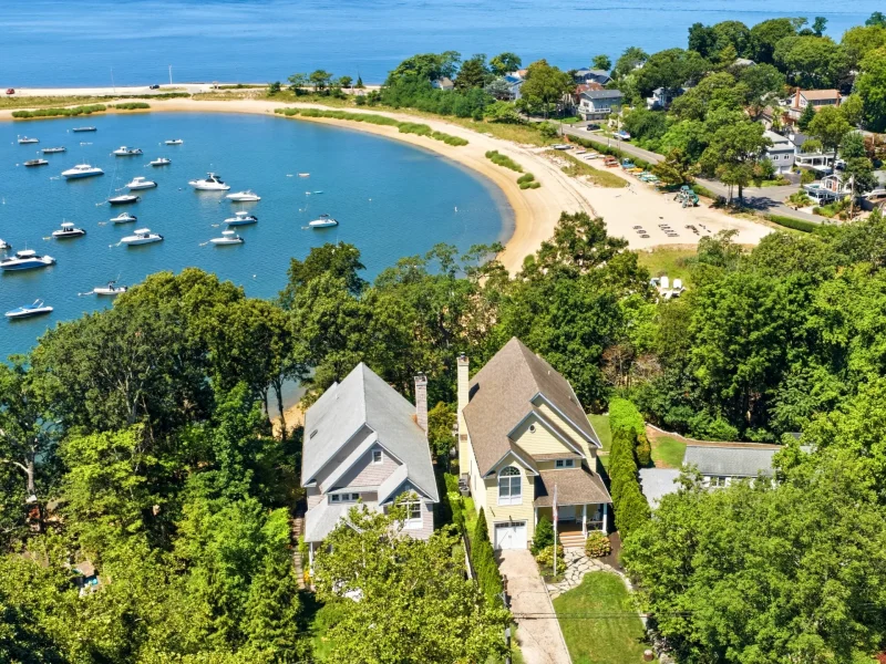 Aerial view of two modern homes surrounded by lush greenery, overlooking a calm bay filled with boats and a sandy beach.