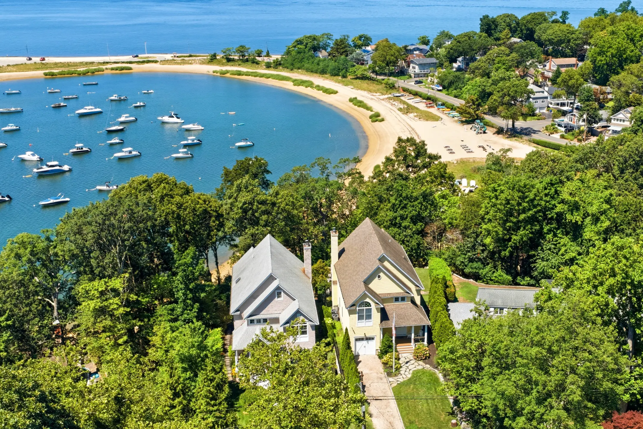 Aerial view of two modern homes surrounded by lush greenery, overlooking a calm bay filled with boats and a sandy beach.