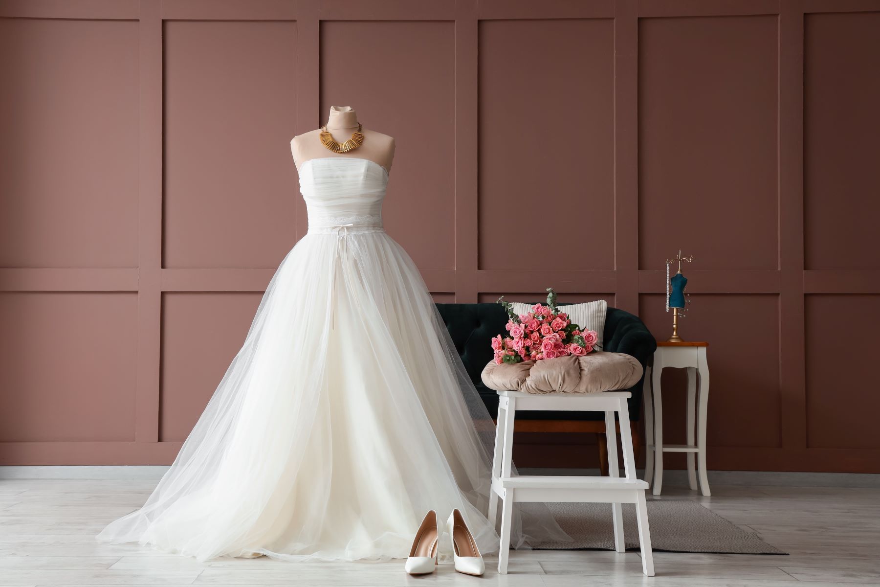 Elegant bridal gown displayed on a mannequin, accompanied by white heels and a bouquet of pink roses in a stylish setting.