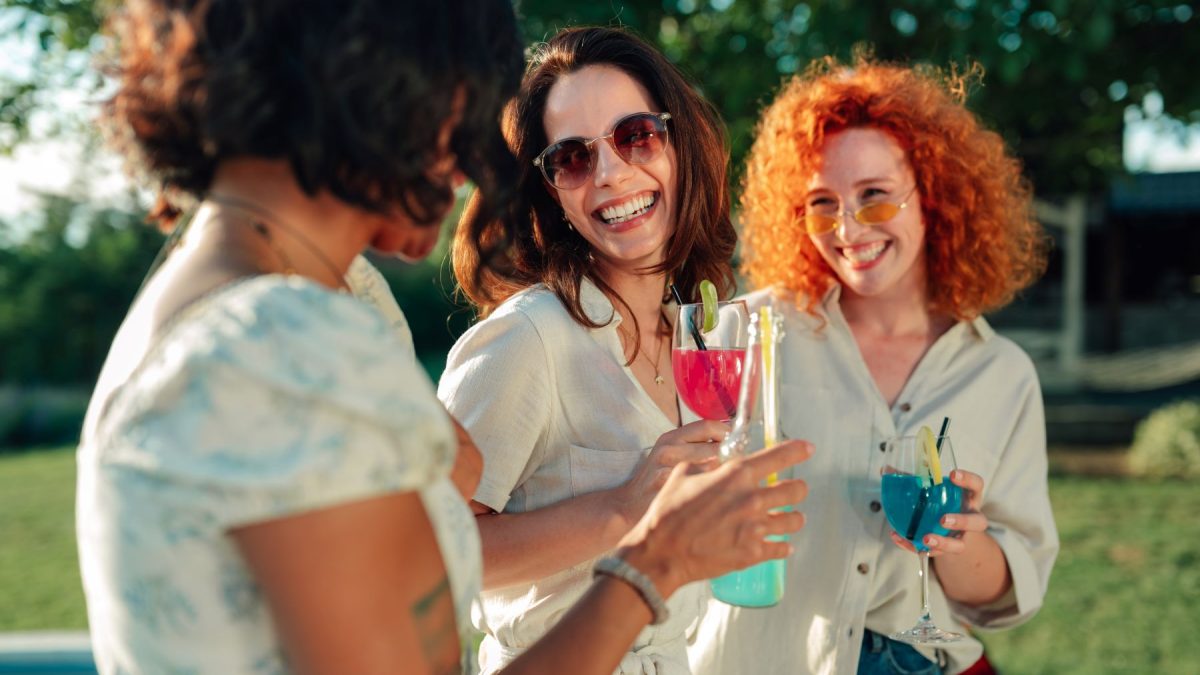Three women enjoy colorful cocktails outdoors, smiling and sharing a joyful moment at a garden party.