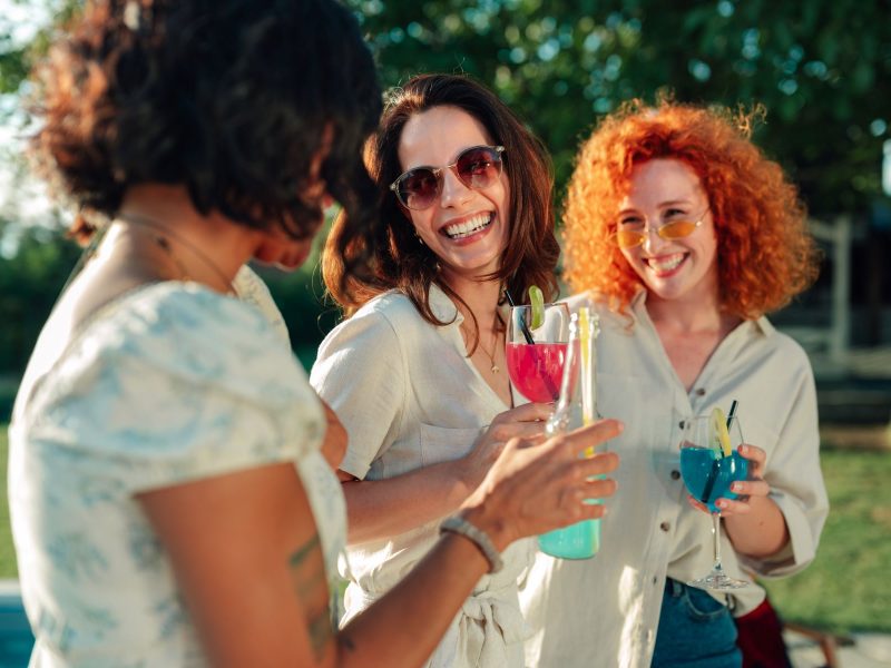 Three women enjoy colorful cocktails outdoors, smiling and sharing a joyful moment at a garden party.