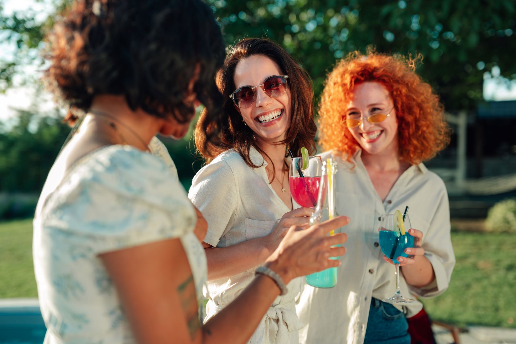 Three women enjoy colorful cocktails outdoors, smiling and sharing a joyful moment at a garden party.