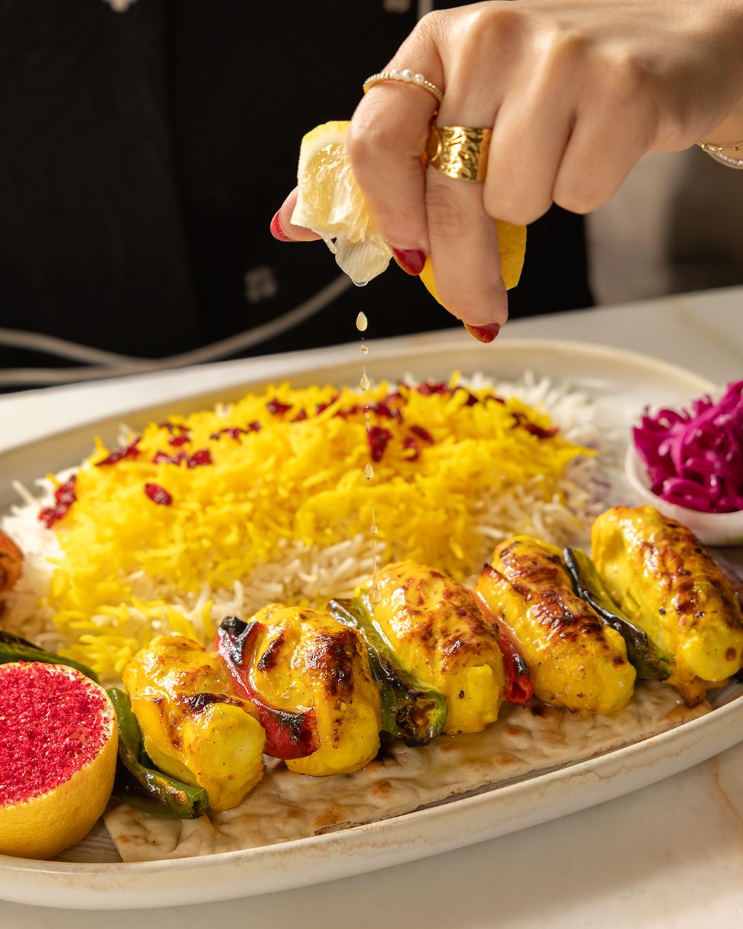 A hand squeezes lemon over a platter of grilled kebabs, saffron rice, and garnishes.