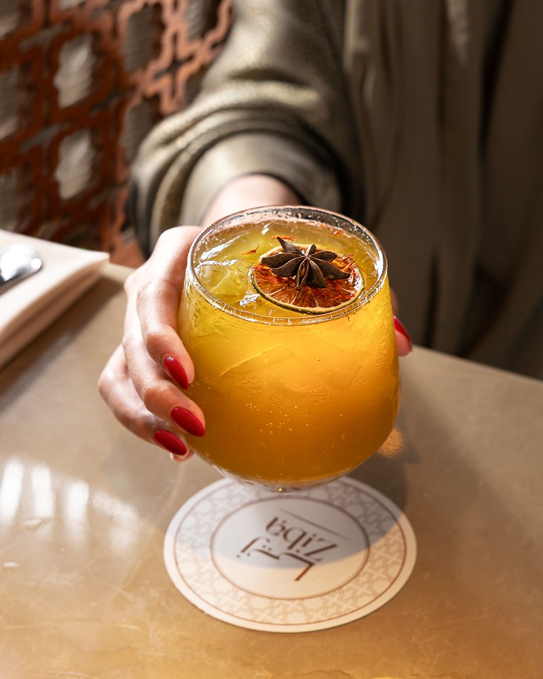 A person holding a glass of yellow beverage garnished with star anise and a slice of dried citrus on a coaster.
