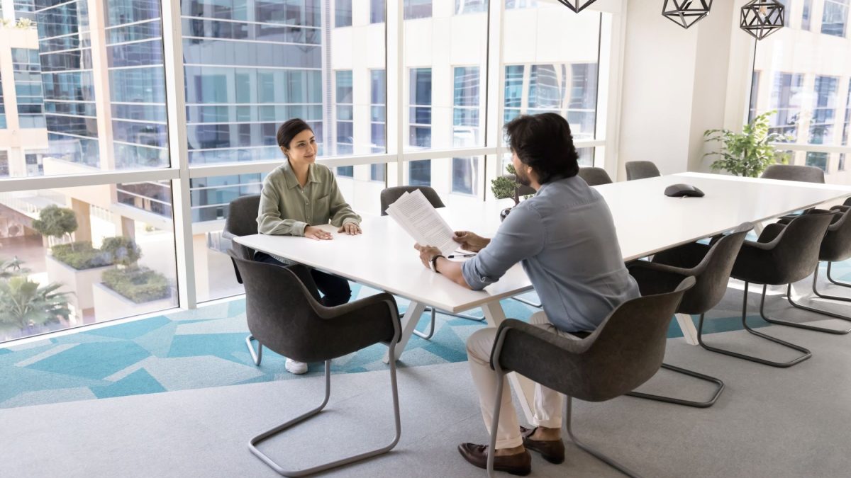 A woman sits confidently during an interview with a man at a modern conference table in a well-lit office space.