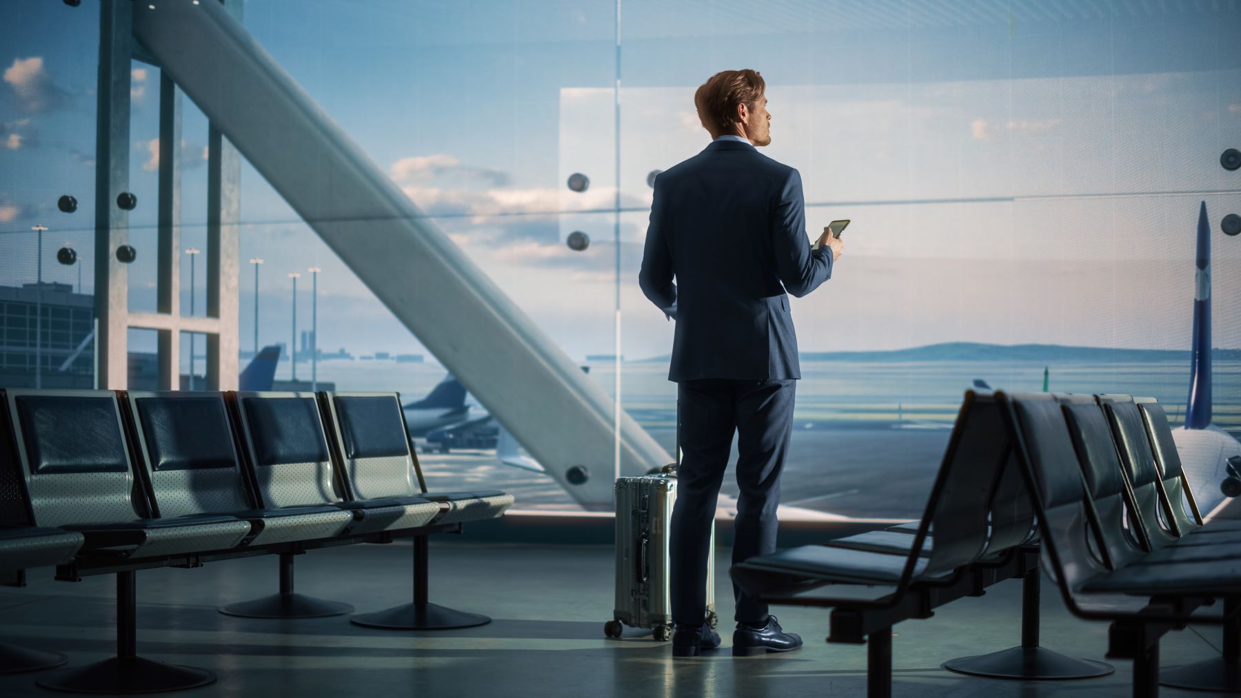 Business traveler waits in an airport terminal, gazing out at the runway while holding a smartphone and a suitcase.