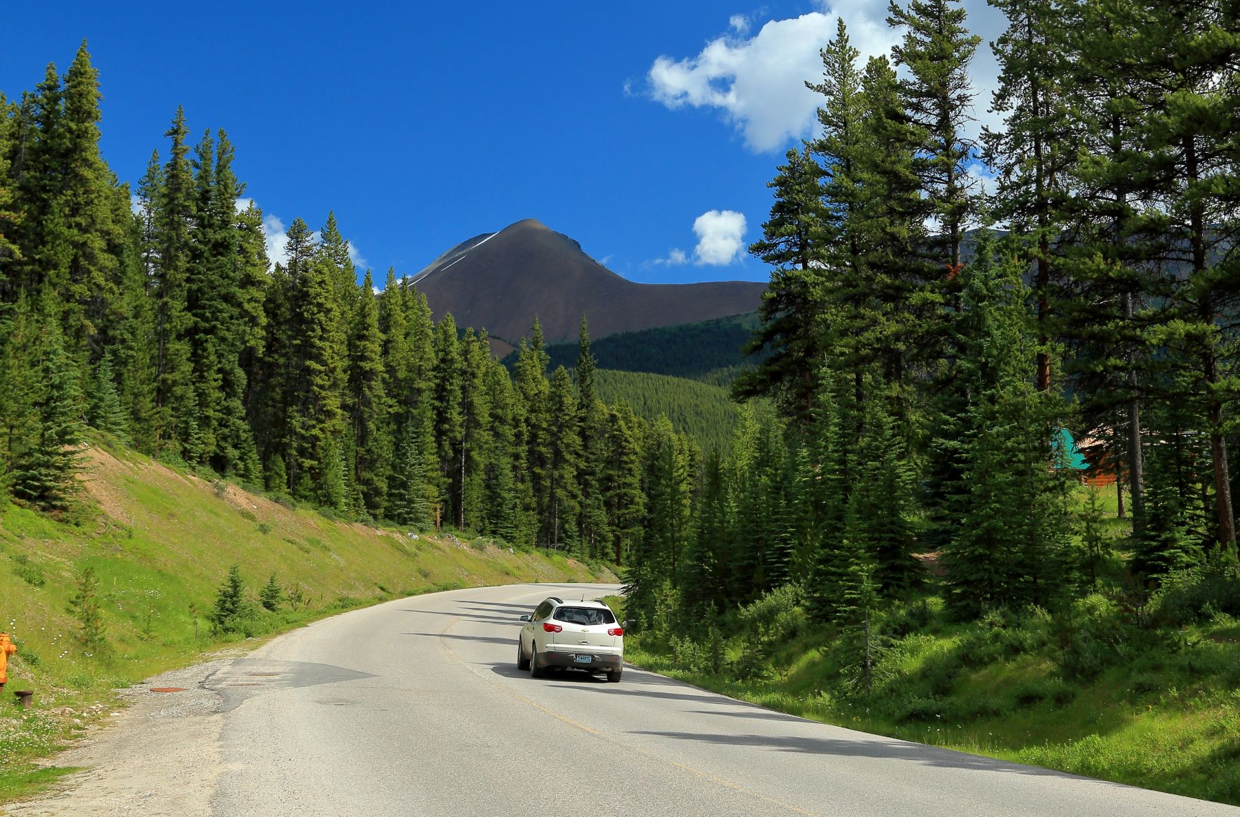 A car drives along a winding two-lane road surrounded by lush green trees and mountains under a clear blue sky.