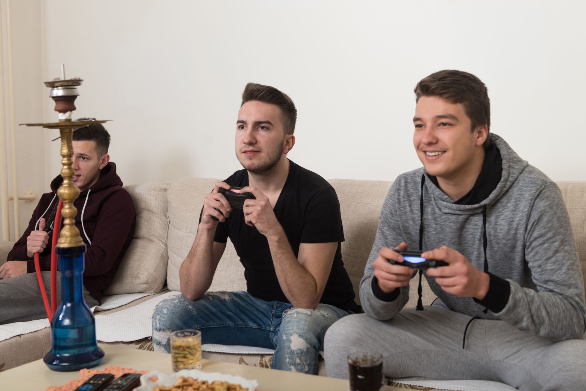Three young men play video games on a sofa with a hookah and snacks on the coffee table, enjoying their time together.