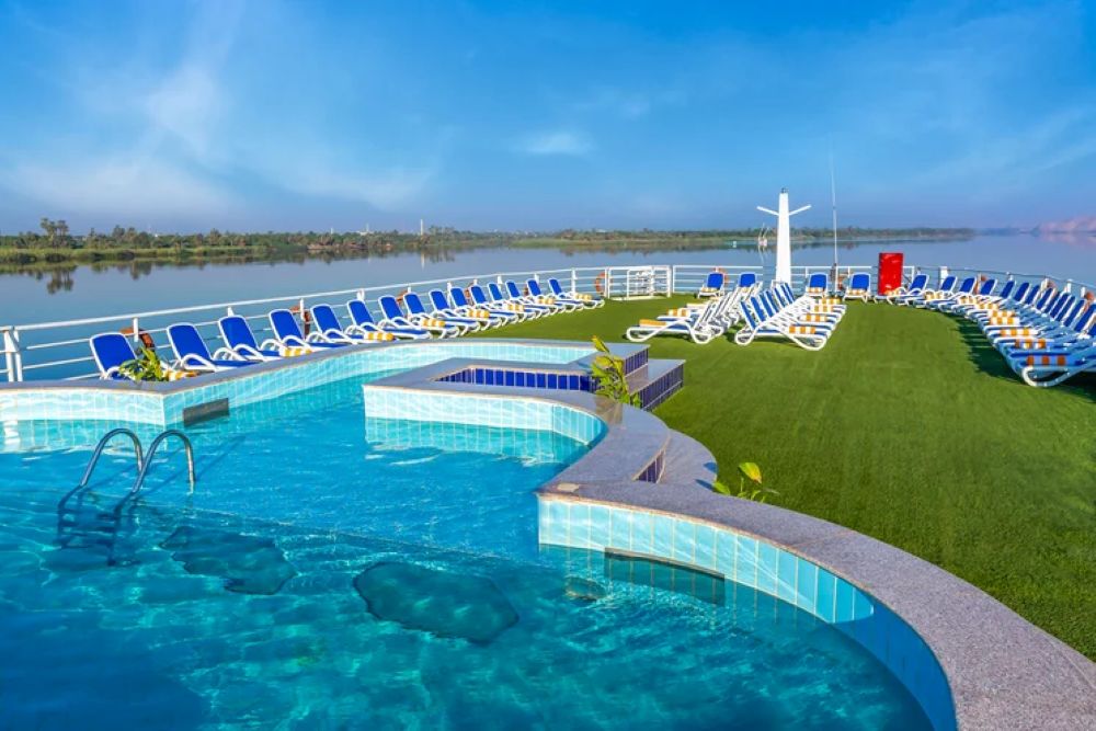 Deck area of a Nile cruise ship featuring a swimming pool and lounge chairs overlooking the river and landscape.