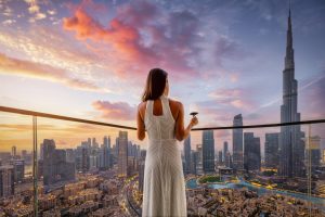 A woman in a white dress enjoys a glass of wine while overlooking a vibrant cityscape at sunset with skyscrapers.