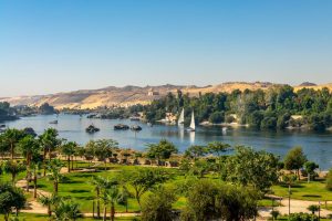 Lush greenery and sailboats line the Nile River, with golden hills in the background under a clear blue sky.