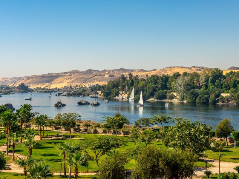 Lush greenery and sailboats line the Nile River, with golden hills in the background under a clear blue sky.