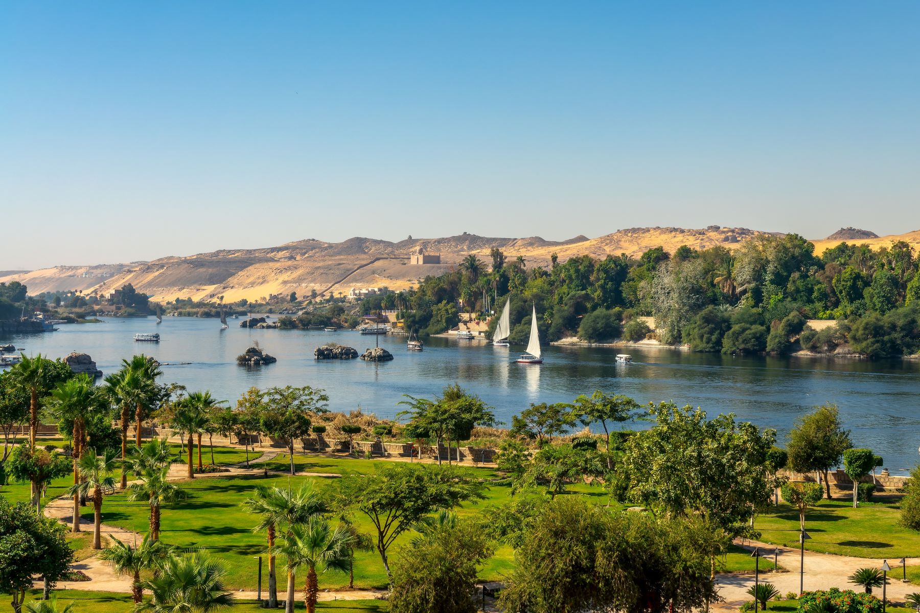 Lush greenery and sailboats line the Nile River, with golden hills in the background under a clear blue sky.