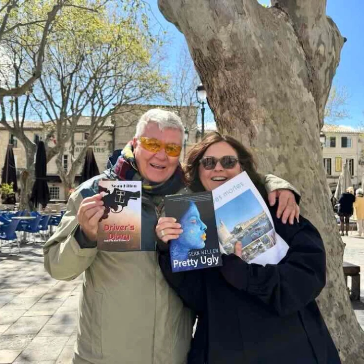 Two people smiling and holding books in a sunlit square, surrounded by trees and outdoor seating in Aigues-Mortes.