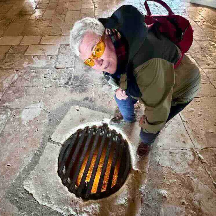 Person kneels by a circular metal grate revealing orange glow, set against the stone floor in a historic building.