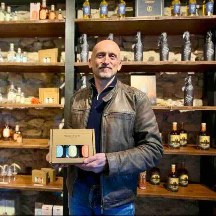 A man holds a gift box with three colorful items in front of shelves filled with various bottles and products.