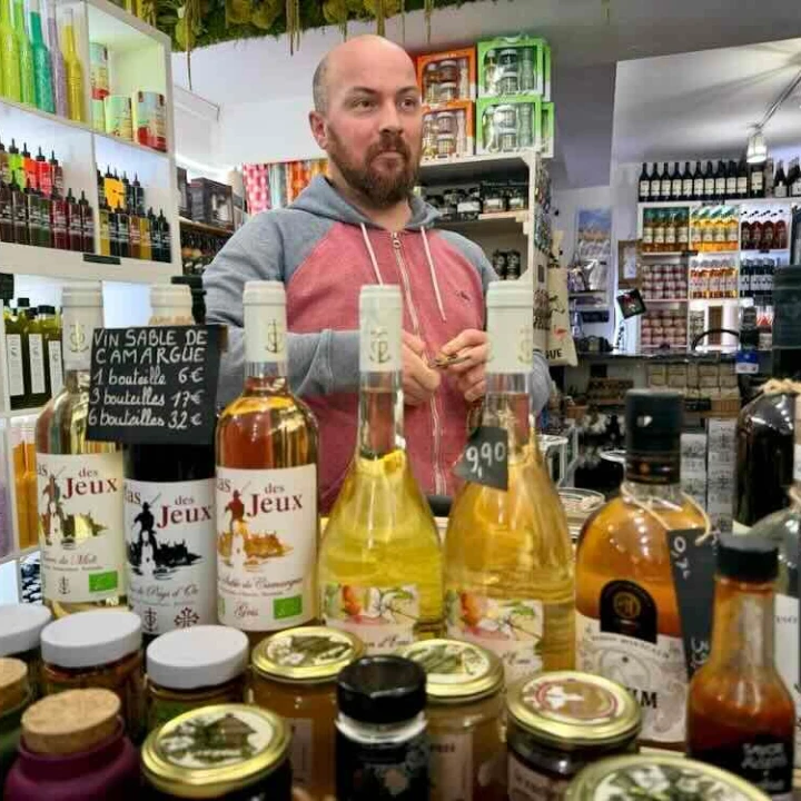 Man standing behind a counter displaying various bottles of wine and jars in a shop in Aigues-Mortes.