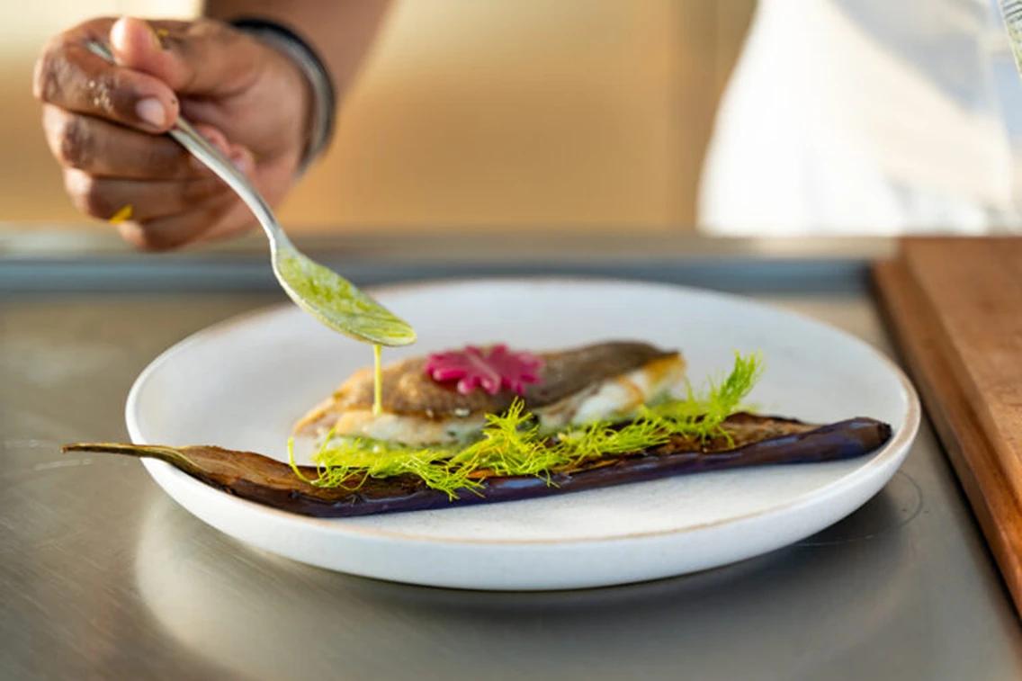 A chef drizzles sauce over a beautifully plated fish dish garnished with greens and a pink flower on a white dish.
