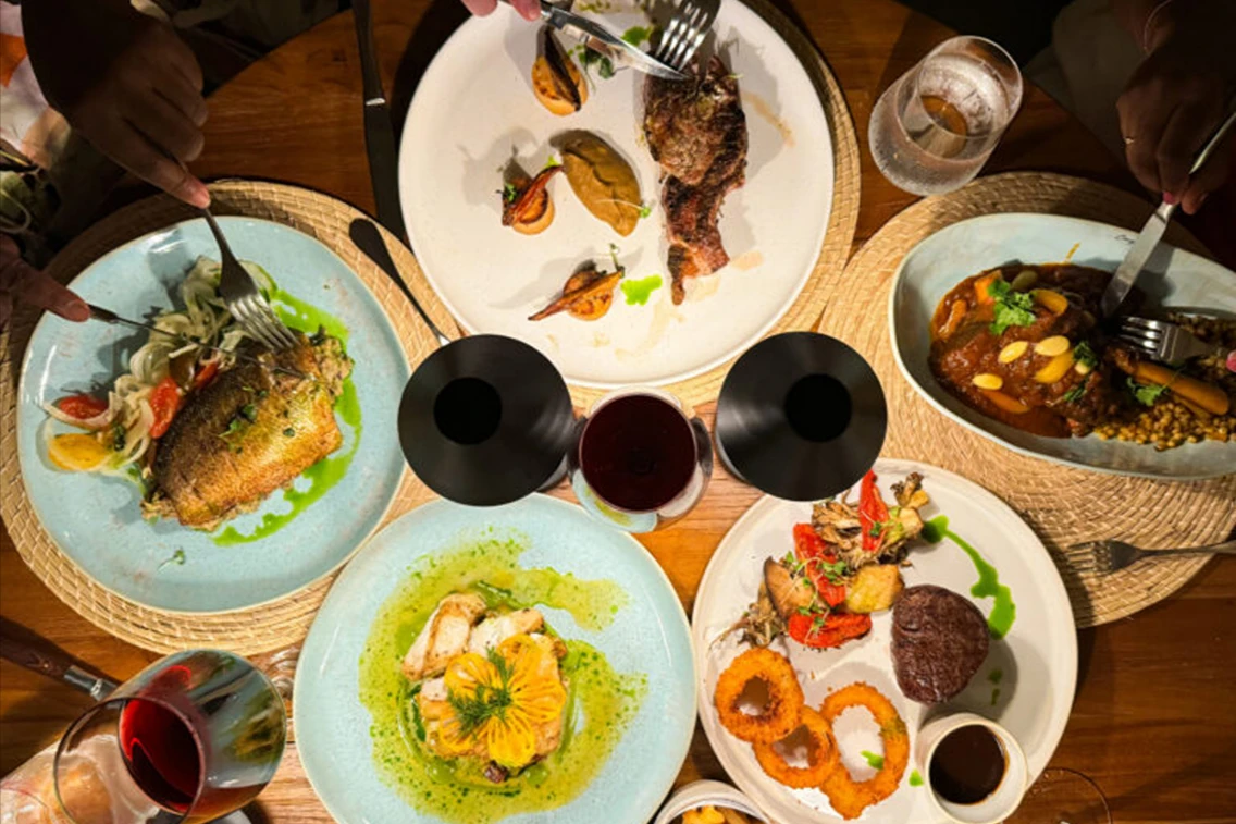 Diverse gourmet dishes are arranged on a table, featuring seafood, meats, and vibrant garnishes, with hands reaching for food.