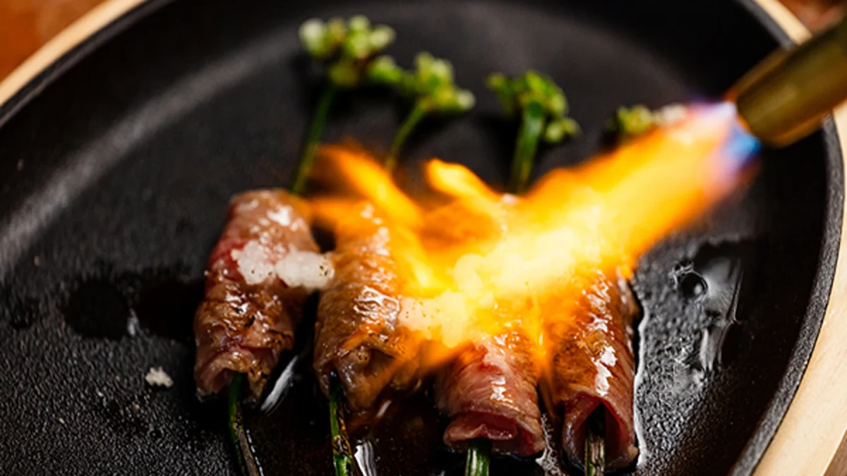 Flame-seared beef rolls wrapped around green onions, served on a sizzling black plate with seasonings.