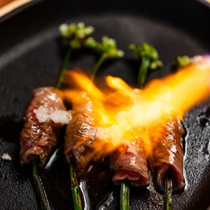 Flame-seared beef rolls wrapped around green onions, served on a sizzling black plate with seasonings.