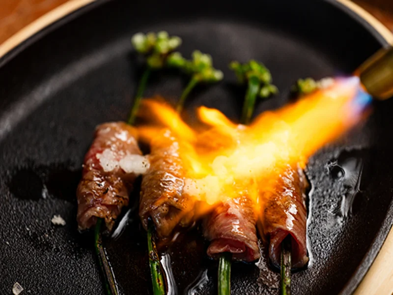 Flame-seared beef rolls wrapped around green onions, served on a sizzling black plate with seasonings.