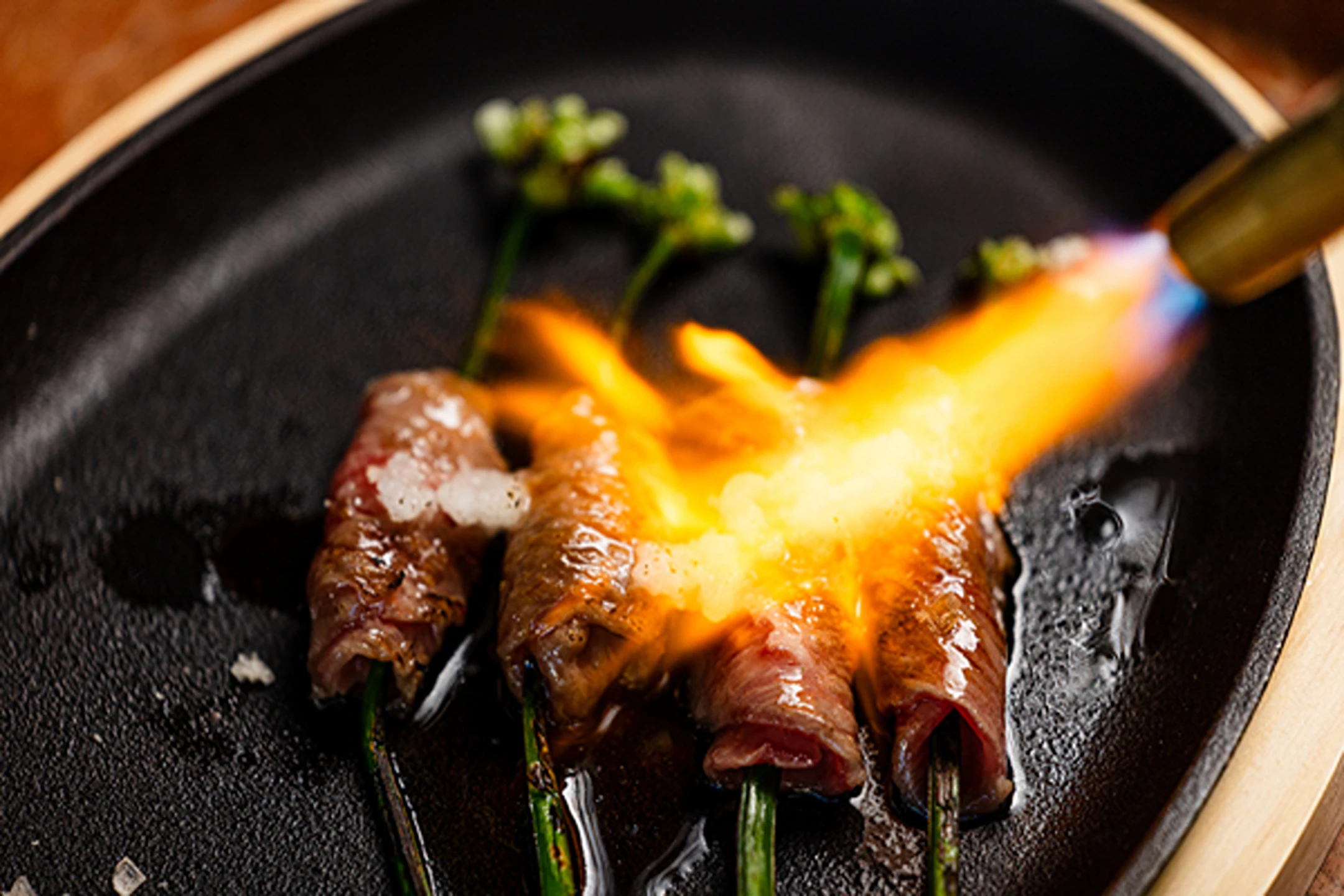 Flame-seared beef rolls wrapped around green onions, served on a sizzling black plate with seasonings.