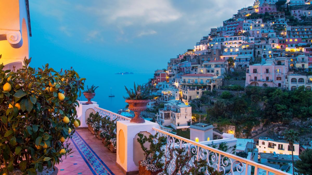 Lemon trees and potted plants line a terrace overlooking the colorful coastal town at twilight in Italy.
