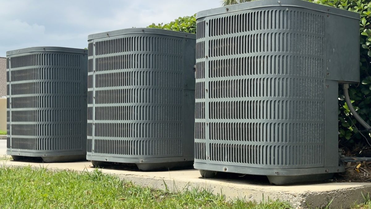 Three air conditioning units installed outdoors on a concrete slab, surrounded by grass and foliage.
