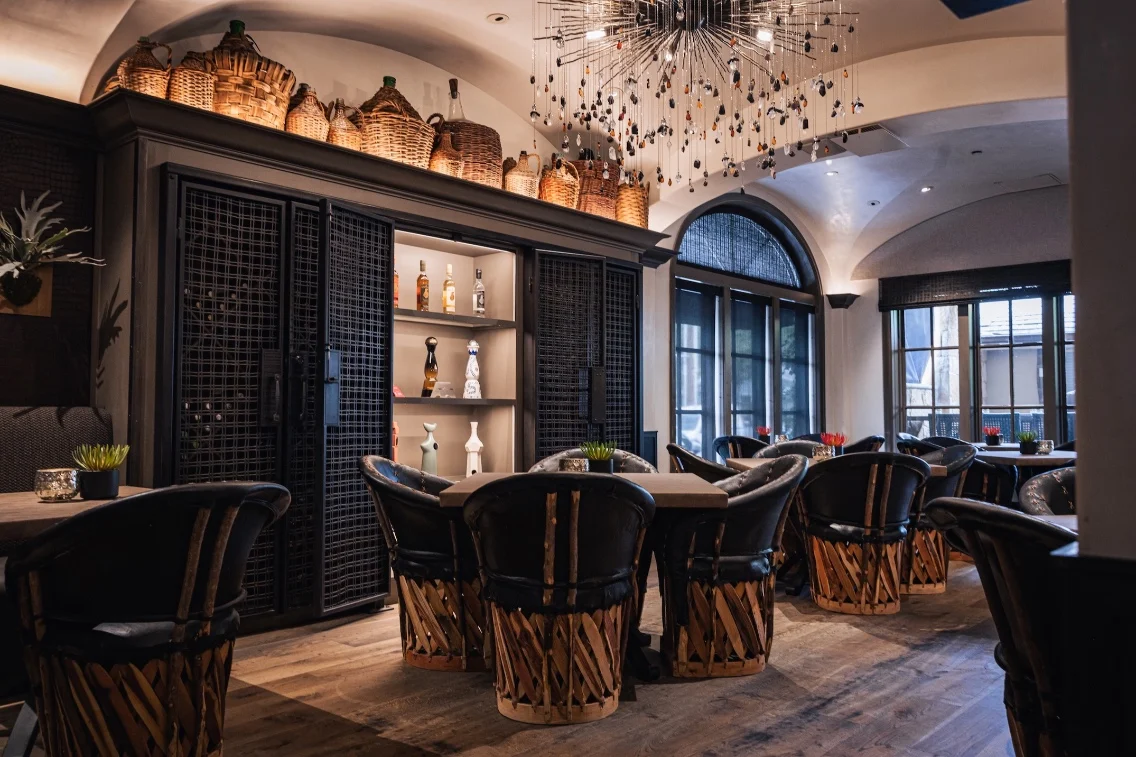Cozy dining area with unique chairs, wooden accents, and decorative shelving displaying bottles and plants.
