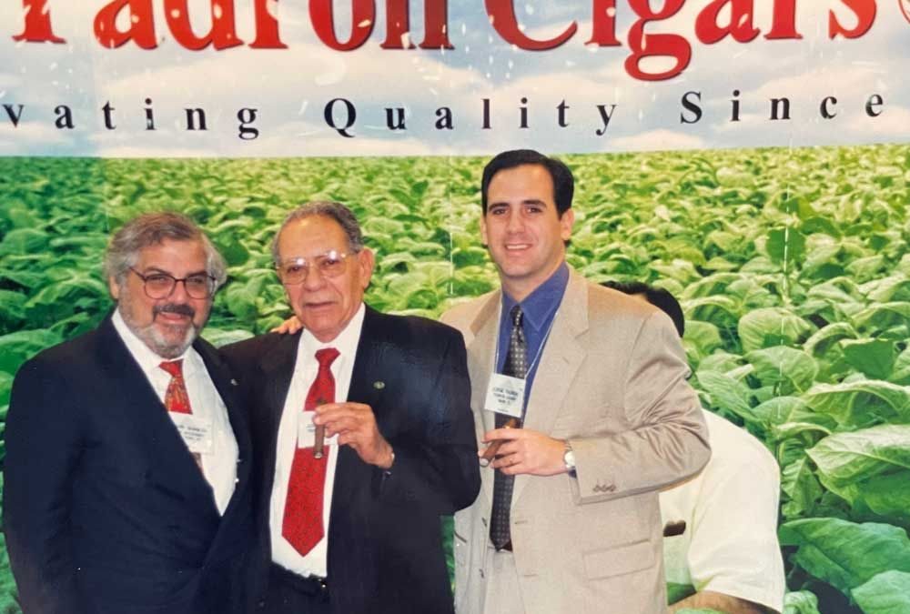 Three men pose together at a Padrón Cigars event, with a crop of tobacco leaves visible in the background.