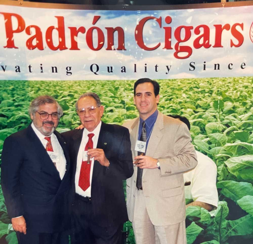 Three men pose together at a Padrón Cigars event, with a crop of tobacco leaves visible in the background.