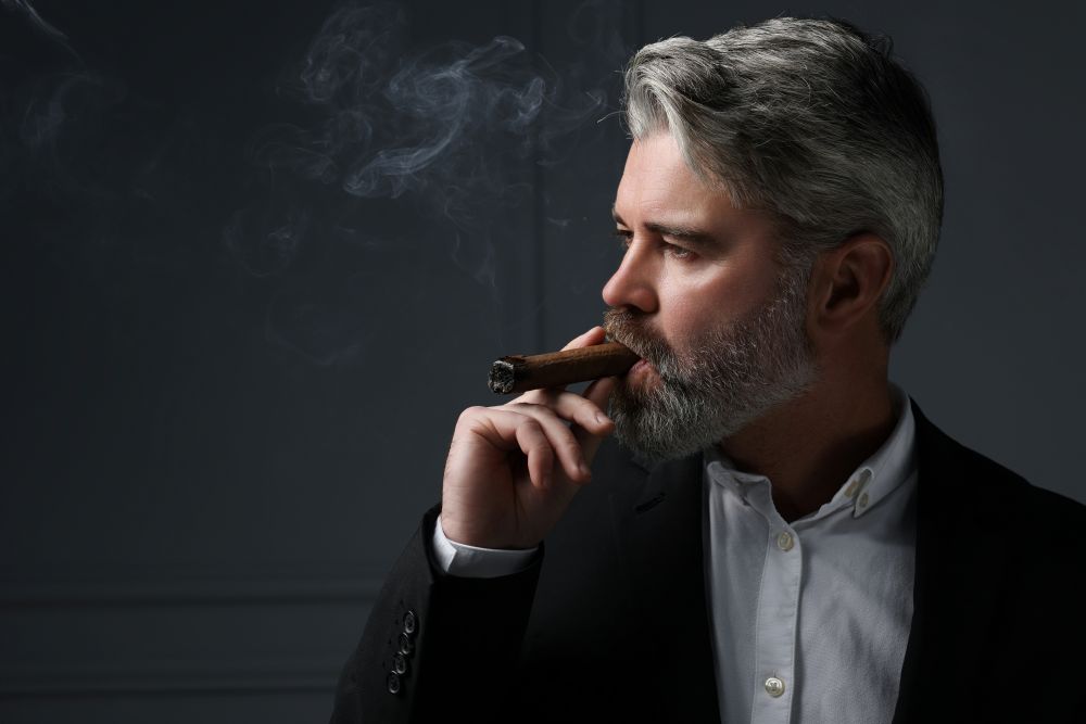 Man in a suit holding a cigar while smoking, with smoke gently billowing in a dimly lit background.