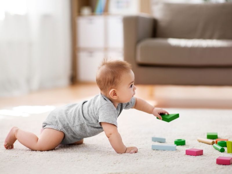Baby crawling on a soft rug, reaching for colorful building blocks scattered around in a bright playroom setting.