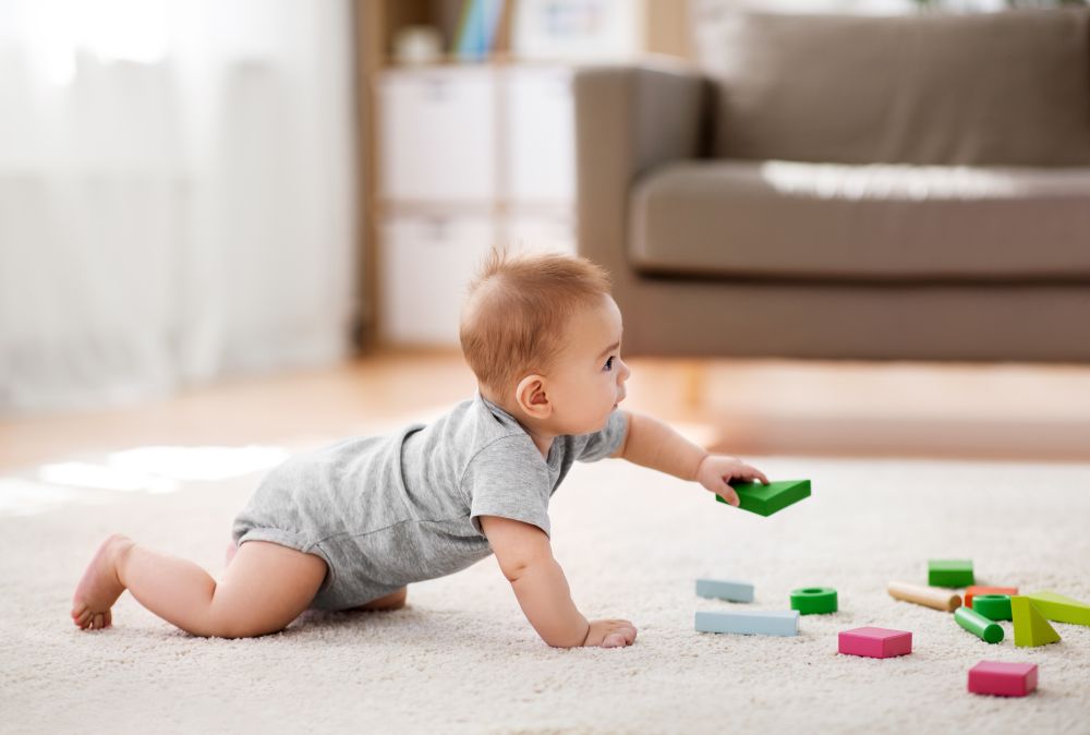 Baby crawling on a soft rug, reaching for colorful building blocks scattered around in a bright playroom setting.