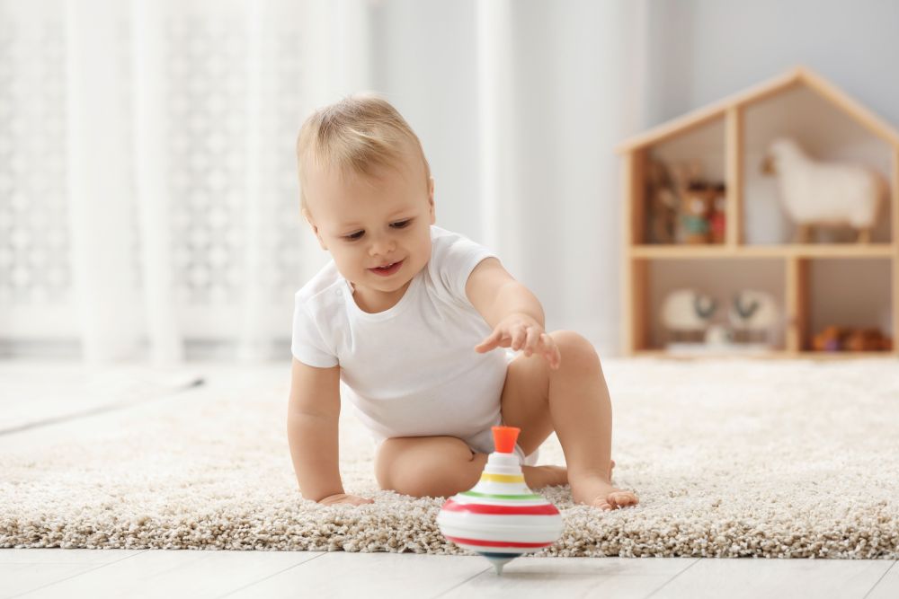 A toddler plays on a soft rug, reaching for a colorful spinning top in a bright and cozy playroom.