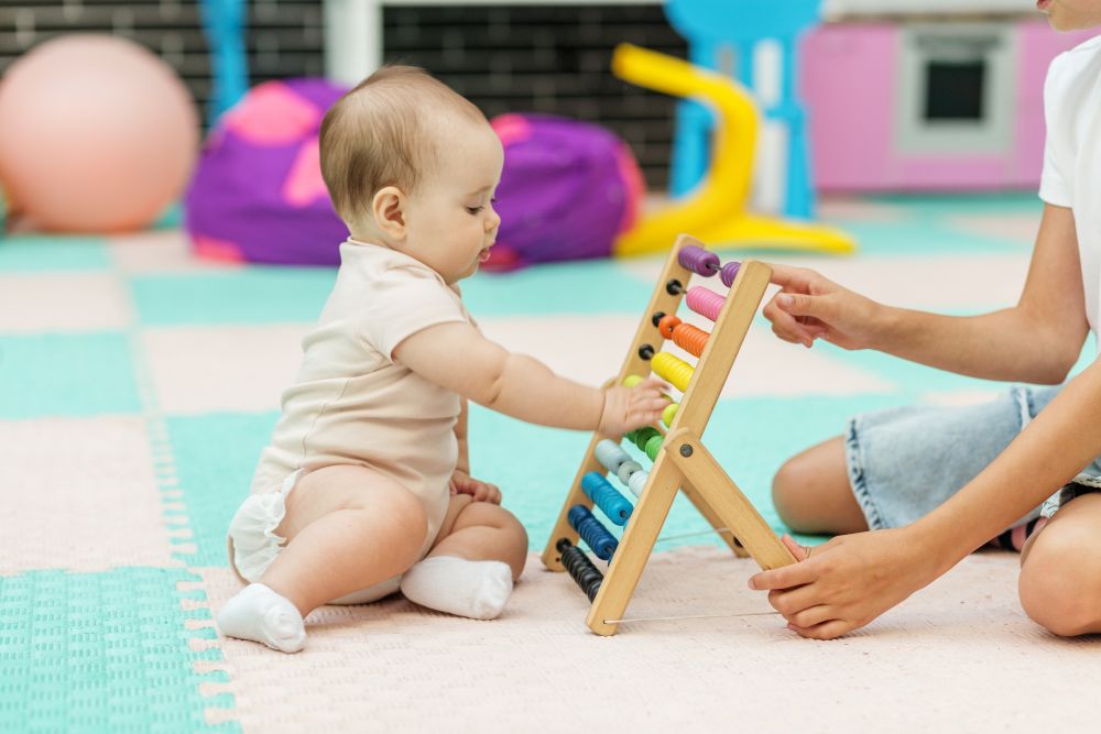 Baby playing with an abacus while an adult guides them in a colorful playroom with soft rugs and mats.