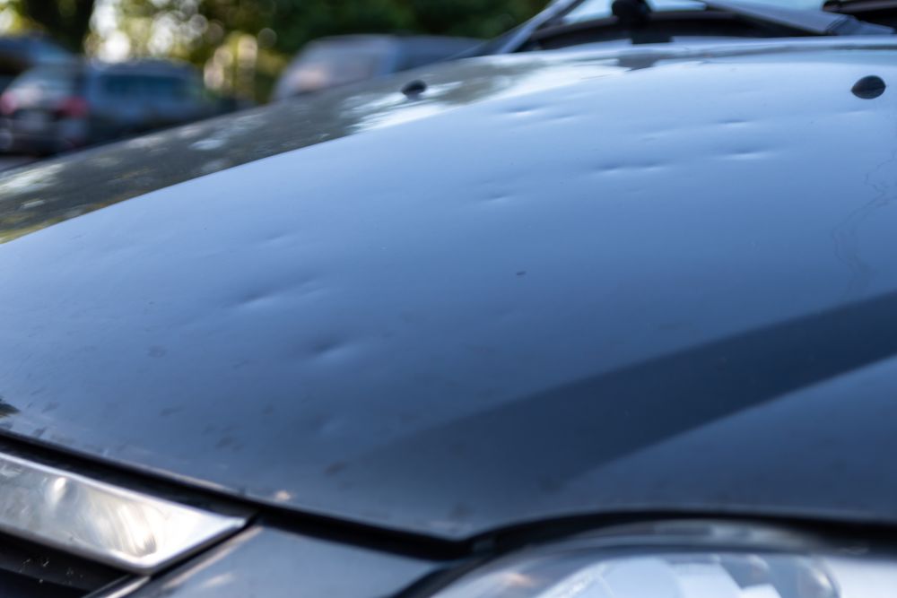 Hood of a black car showing multiple dents, likely caused by hail damage, with blurred vehicles in the background.