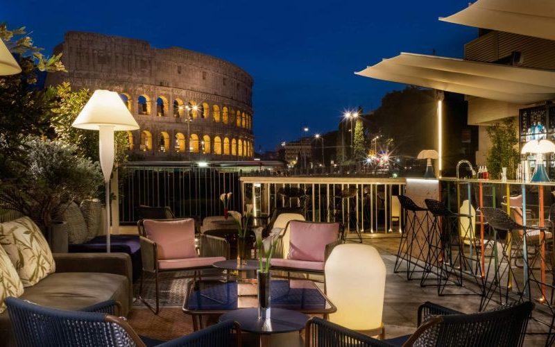 A stylish outdoor seating area with modern furniture overlooking the Colosseum, illuminated at night with warm lighting.