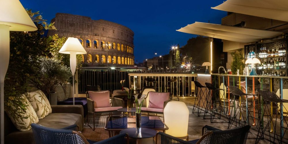 A stylish outdoor seating area with modern furniture overlooking the Colosseum, illuminated at night with warm lighting.