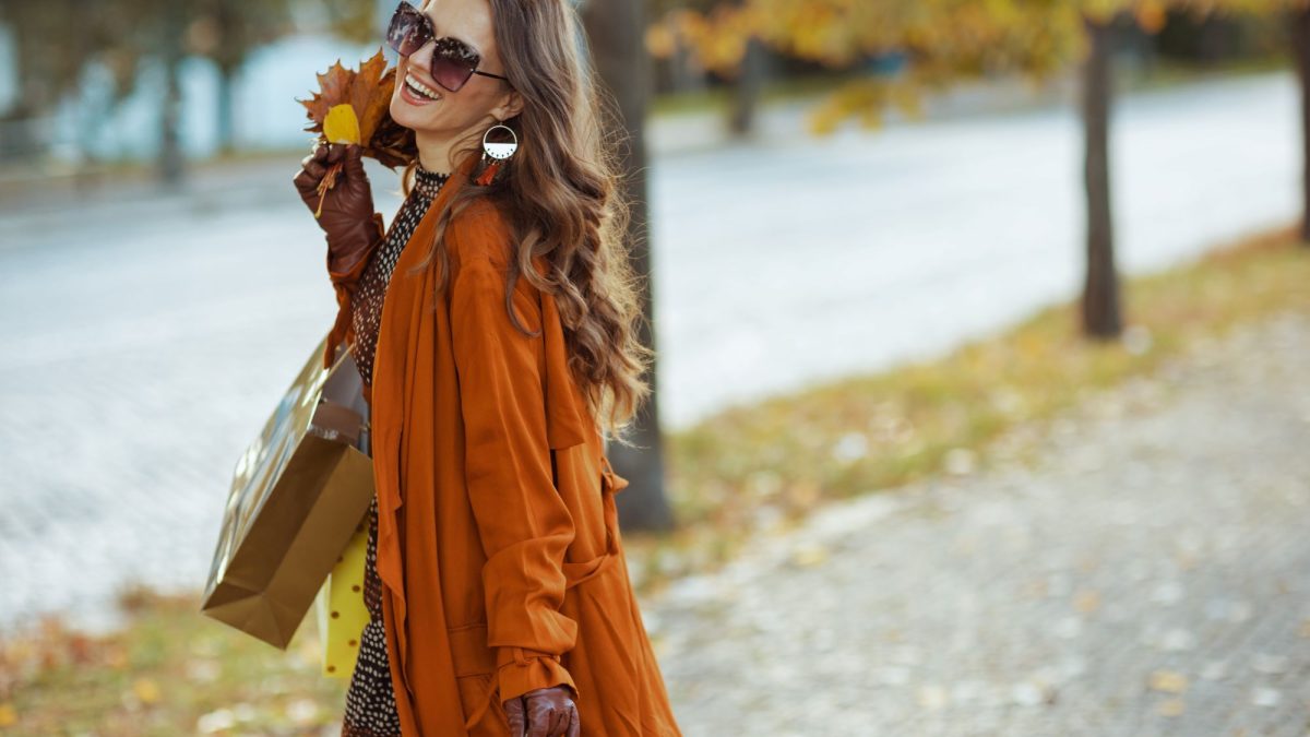 Woman in an orange coat holding shopping bags and autumn leaves, smiling while walking along a tree-lined street.