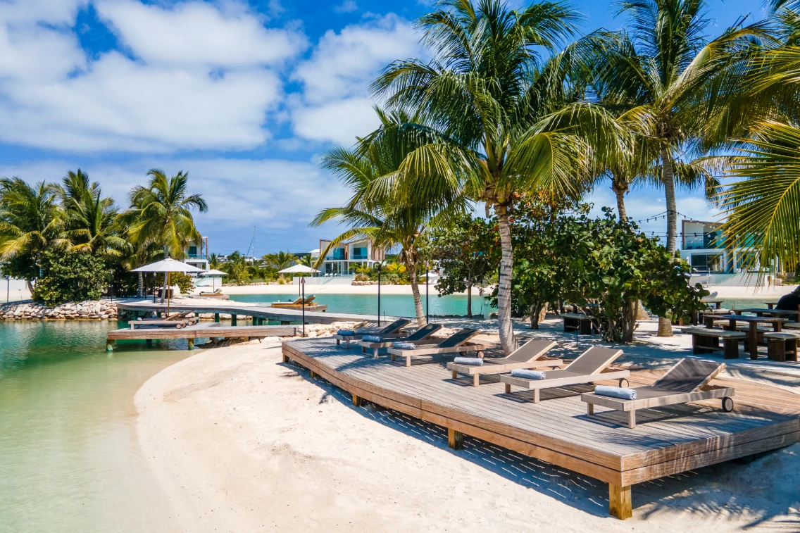 Luxurious wooden deck chairs beside a calm turquoise lagoon, surrounded by palm trees and tropical landscaping in Turks & Caicos.