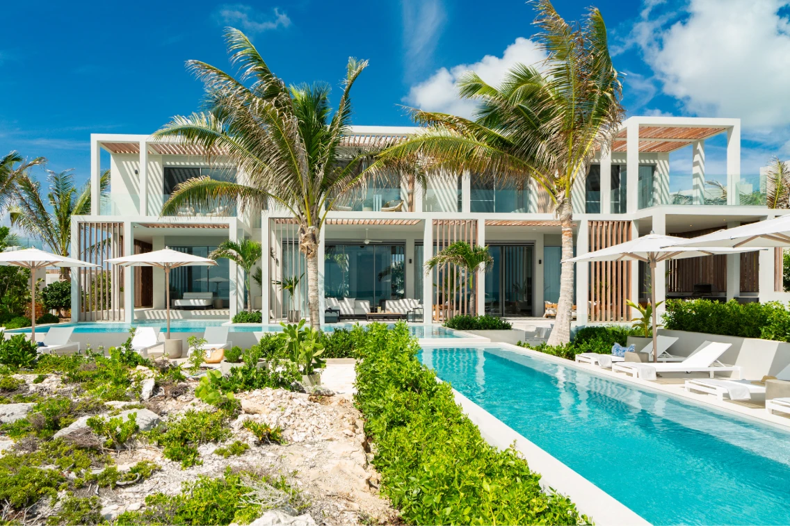Modern beachfront villa featuring lush greenery, a swimming pool, and relaxing areas under umbrellas.