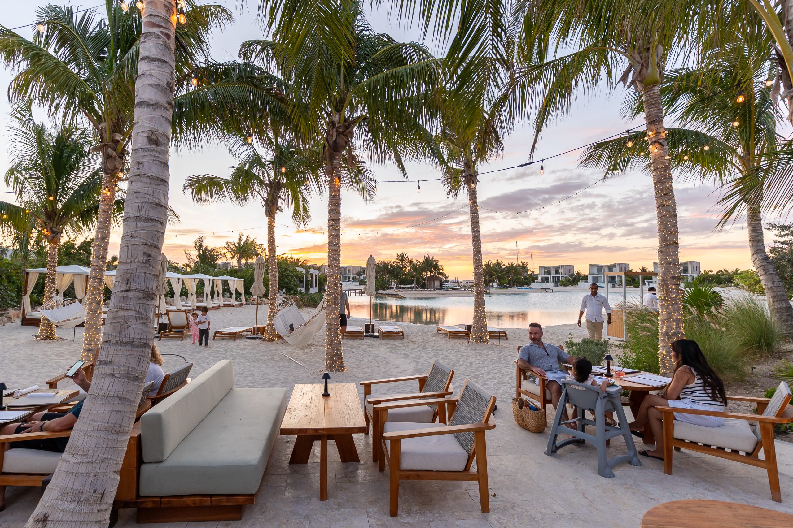 Lush palm trees frame a serene sand area with seating, hammocks, and a waterfront view at sunset in Turks & Caicos.
