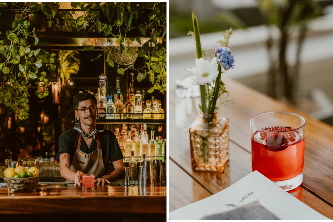 Bartender at Acre Resort prepares drinks with a backdrop of plants and shelves filled with spirits, alongside a cocktail and flower vase.