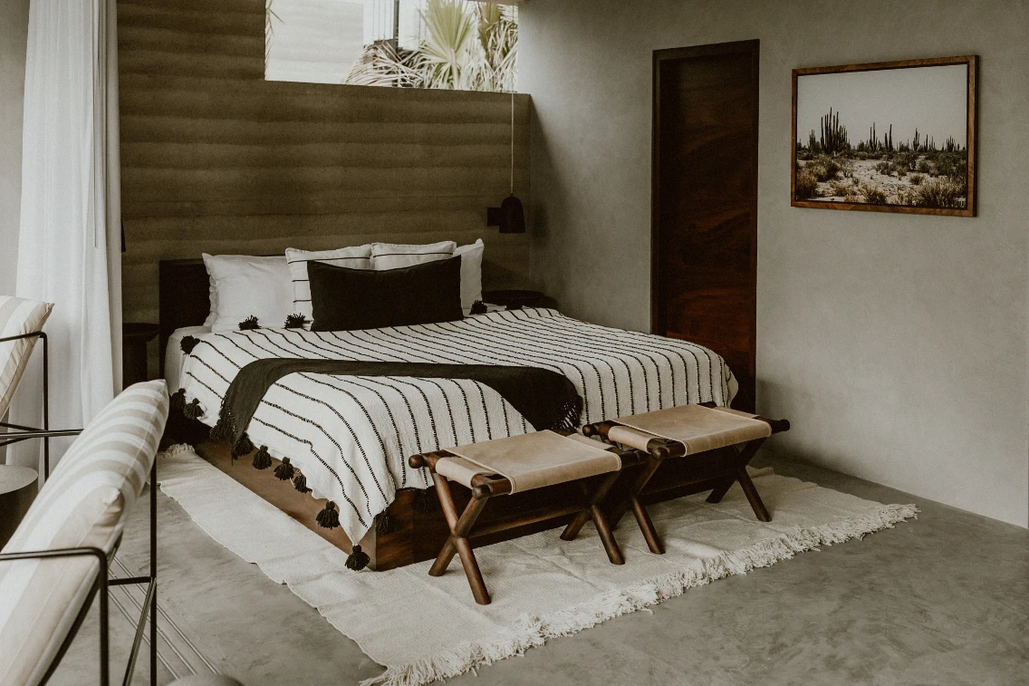 Modern bedroom featuring a striped bedspread, minimalist decor, and earthy tones, with a desert landscape artwork on the wall.